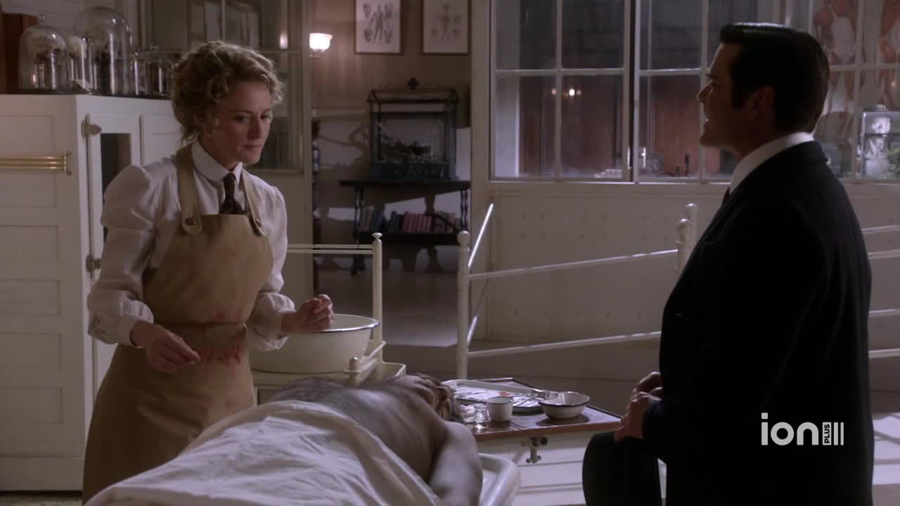 A woman in a blood-stained apron stands over a body on a table, gesturing with her hands. A man in a dark suit watches her intently.

