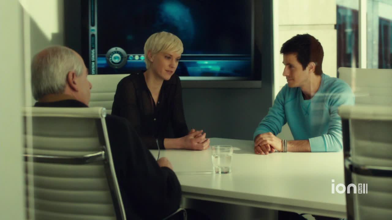 Three people sit around a table, their hands resting on the white surface. The woman in black looks toward the man in the blue sweater, while the older man faces forward.
