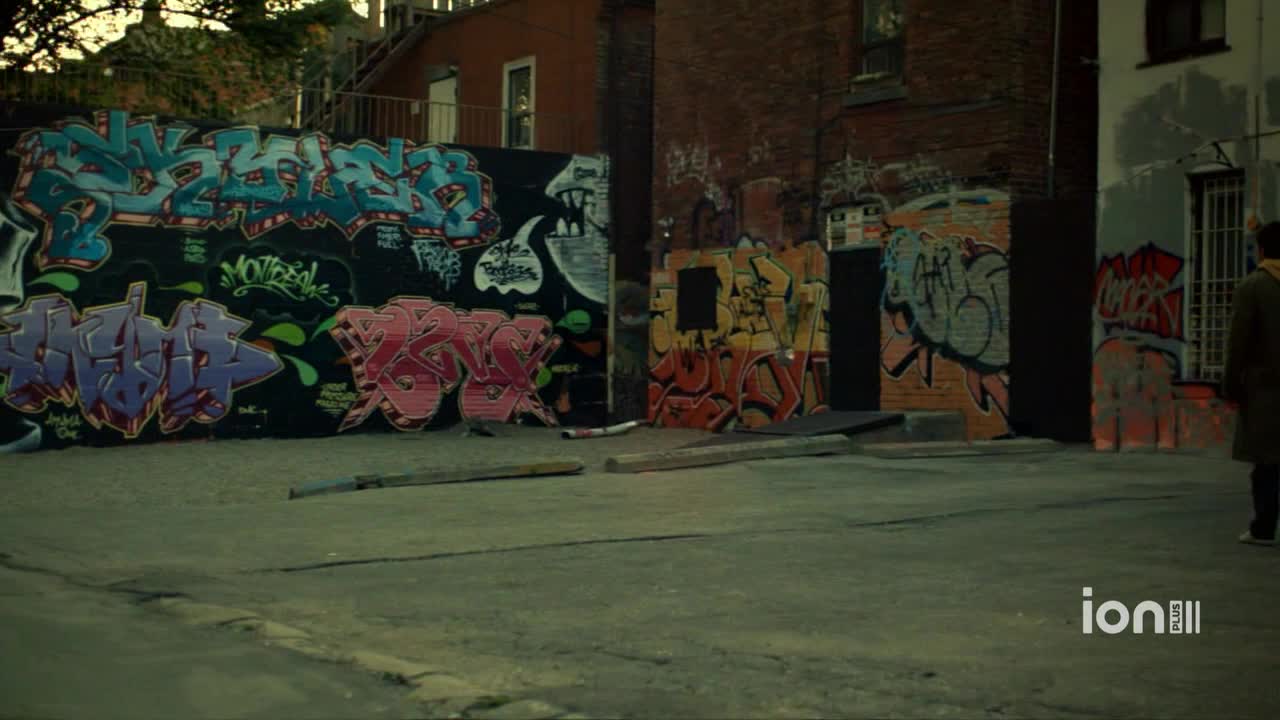 Graffiti covers the brick walls and a dark panel in an alley. A person in a long coat walks away from the camera, toward the right side of the frame.
