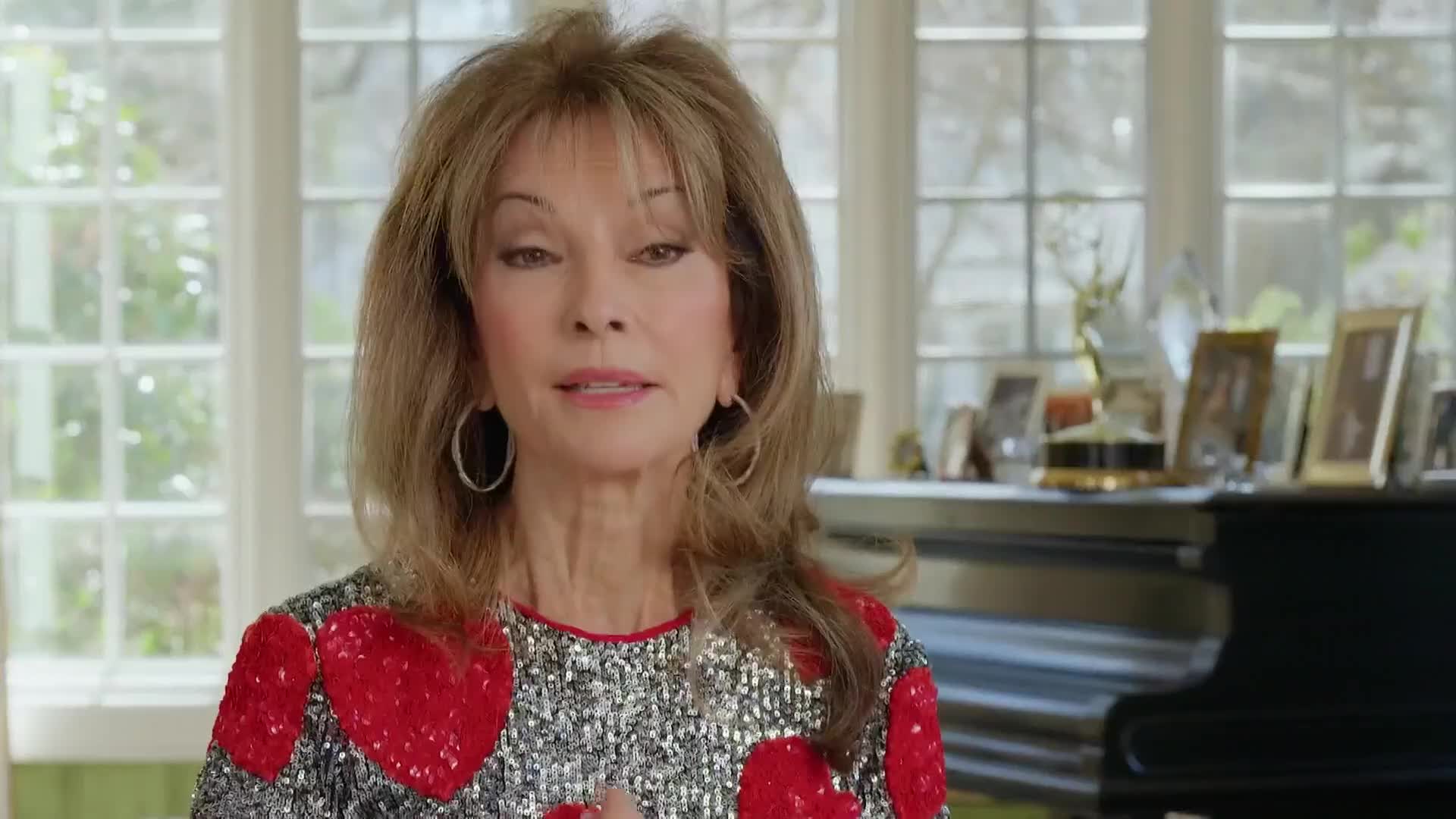 A woman with long, light brown hair and silver hoop earrings is speaking, her lips moving as she talks. She's wearing a sequined top with red heart shapes, and a piano sits behind her.
