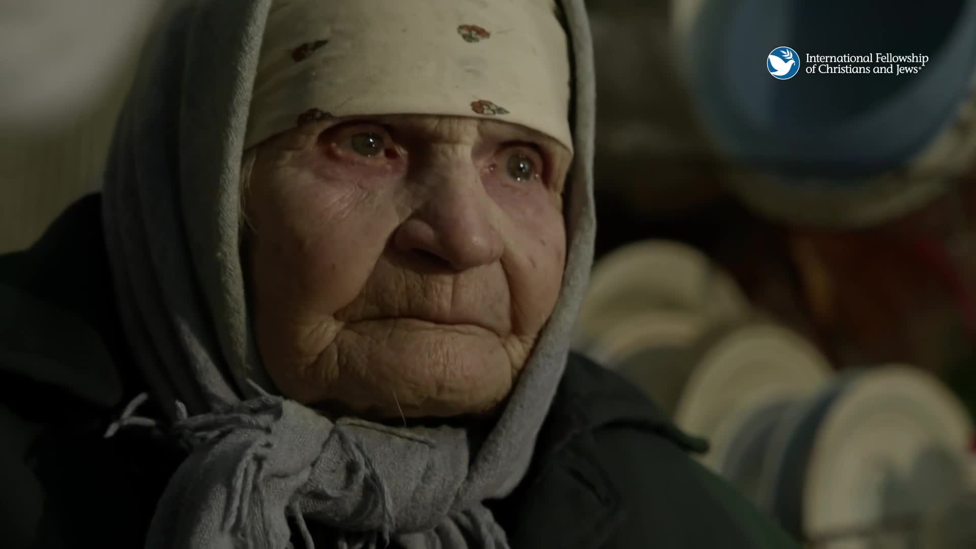 An elderly woman, her face etched with deep lines, looks directly ahead. She wears a headscarf and a grey shawl, her eyes reflecting a quiet strength.