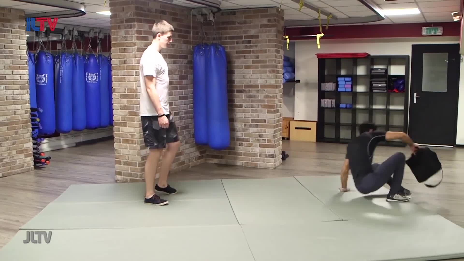 A man in a white t-shirt stands near a row of blue punching bags. Across the room, another man is on the floor, mid-movement, with a black bag beside him.