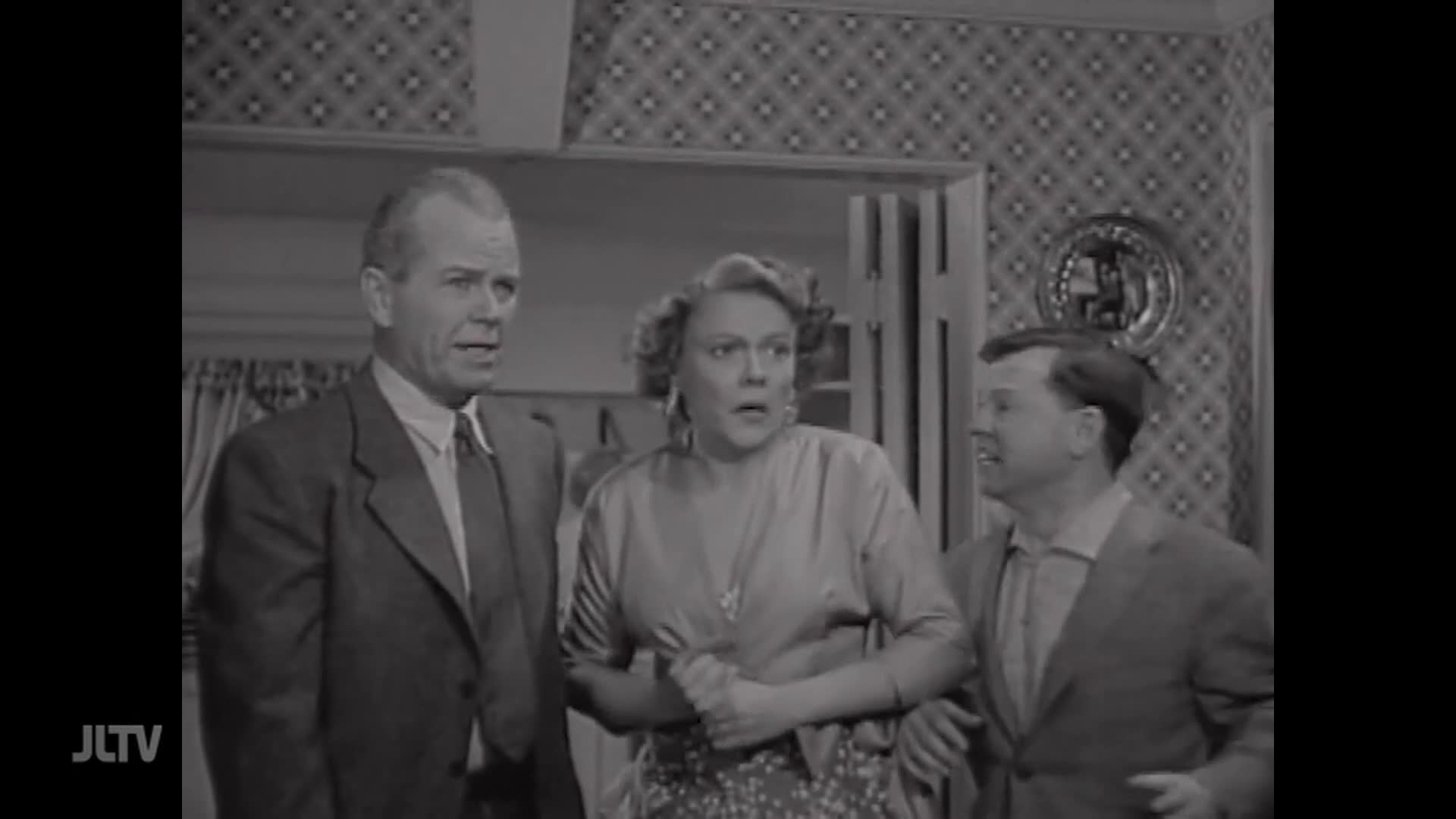A man in a suit and tie stands to the left, his brow furrowed. A woman in a patterned dress and a man in a collared shirt and jacket are beside him, their expressions a mix of surprise and concern. The JTLV logo is visible in the bottom left corner.
