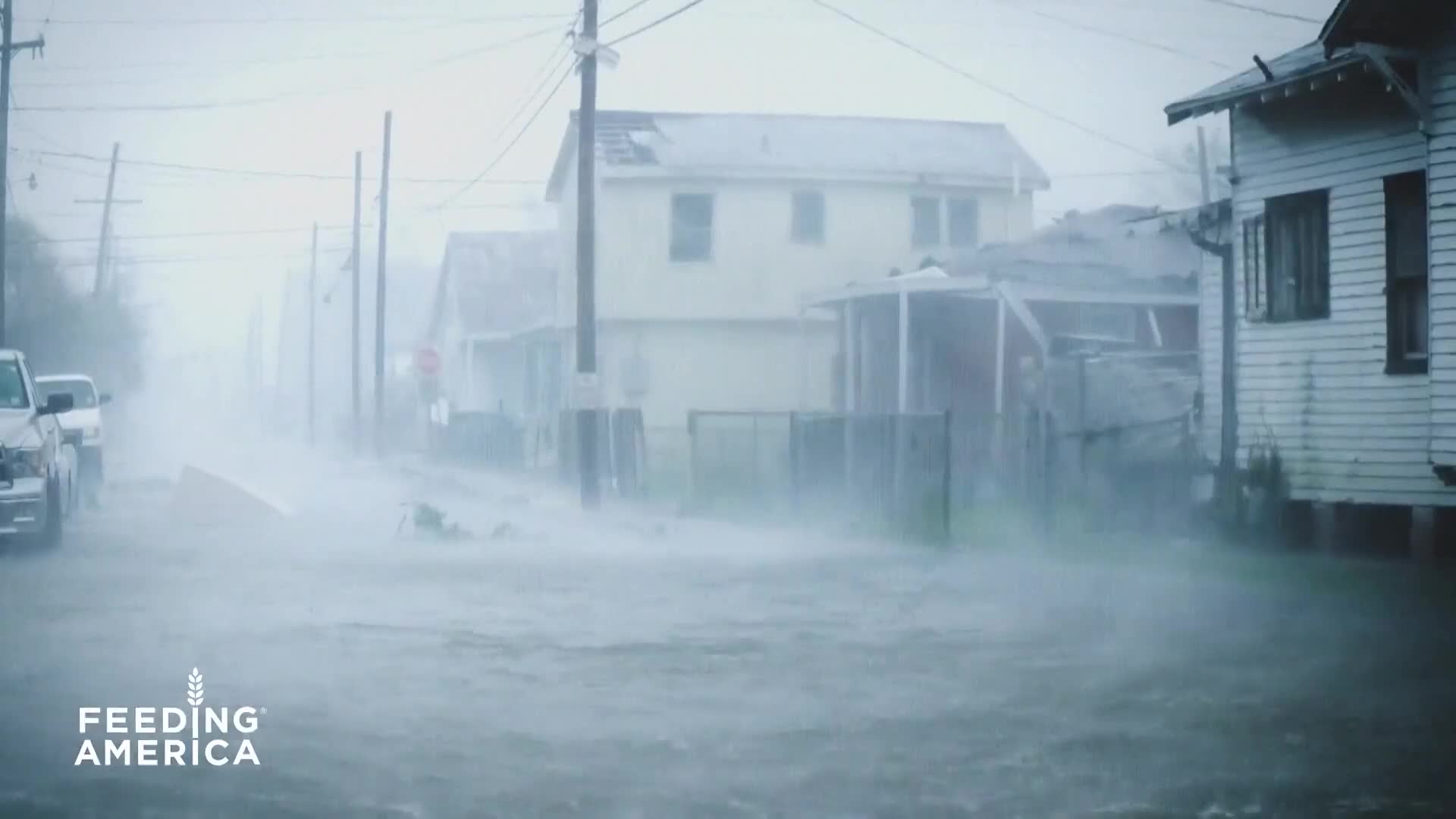 The street is a churning mess of water and debris, with houses on either side battered by wind and rain. A white truck sits partially submerged, its tires disappearing into the rising flood.
