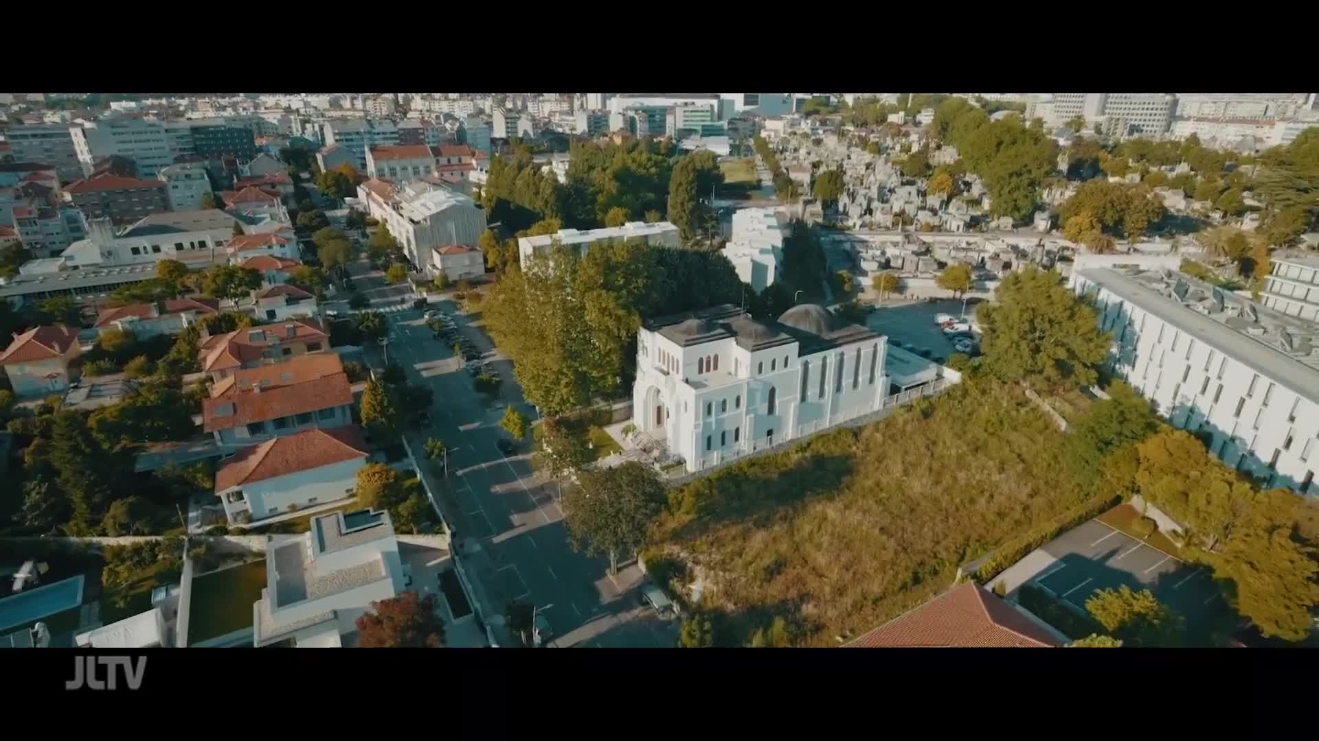 A white building with a domed roof stands prominently in the center, surrounded by trees and a street. Beyond it, rows of gravestones stretch across a hillside, a quiet testament to lives lived.