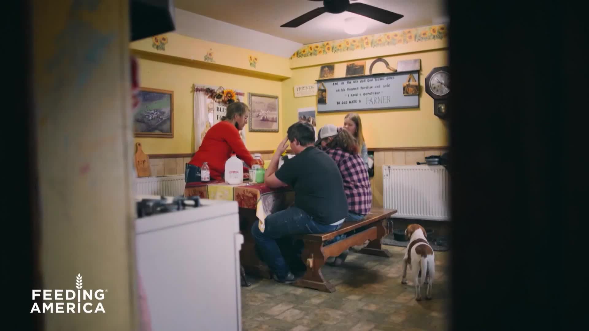 A woman in a red shirt pours from a jug on a table where a family sits. A small dog stands by the bench, watching.