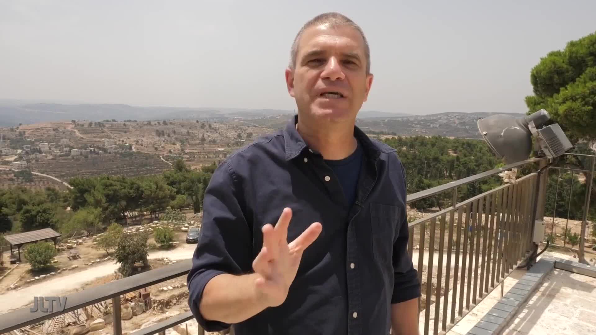A man in a dark blue shirt gestures with his left hand while speaking. Behind him, a sprawling landscape of hills and scattered buildings stretches under a hazy sky.