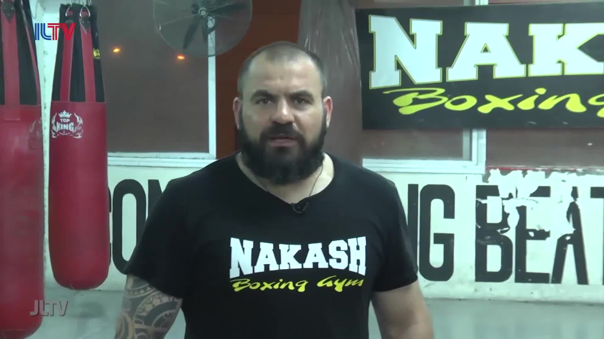 A man with a beard stands in front of hanging red punching bags. He wears a black t-shirt that reads "Nakash Boxing Gym."