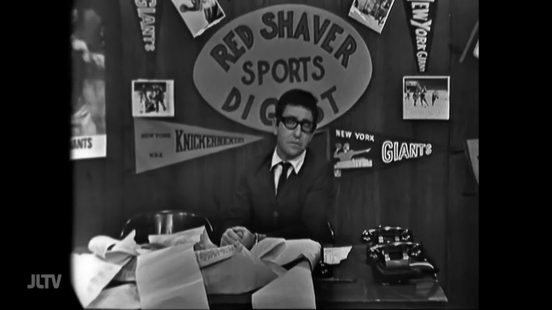 A man in a suit and glasses sits at a desk, looking directly ahead. Pennants for the New York Giants and Knickerbockers hang behind him, along with a sign for "Red Shaver Sports Digest."