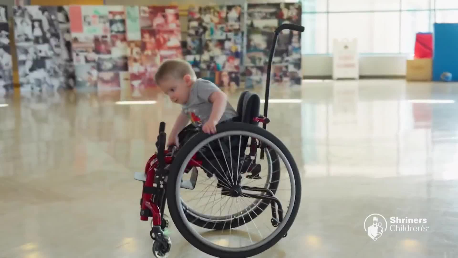A young boy in a wheelchair reaches for the large rear wheel. He's in a bright, open space with a wall of photos behind him.