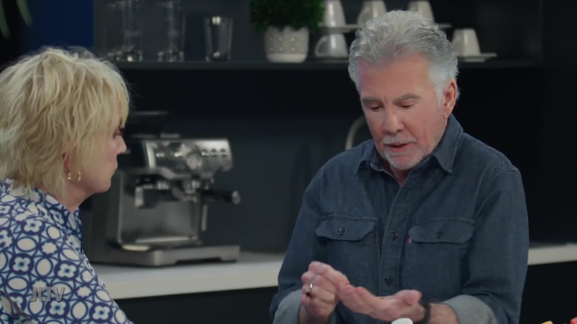 A man with graying hair gestures with his hands while speaking, his denim shirt buttoned. Beside him, a woman with blonde hair looks on, her patterned blouse visible.