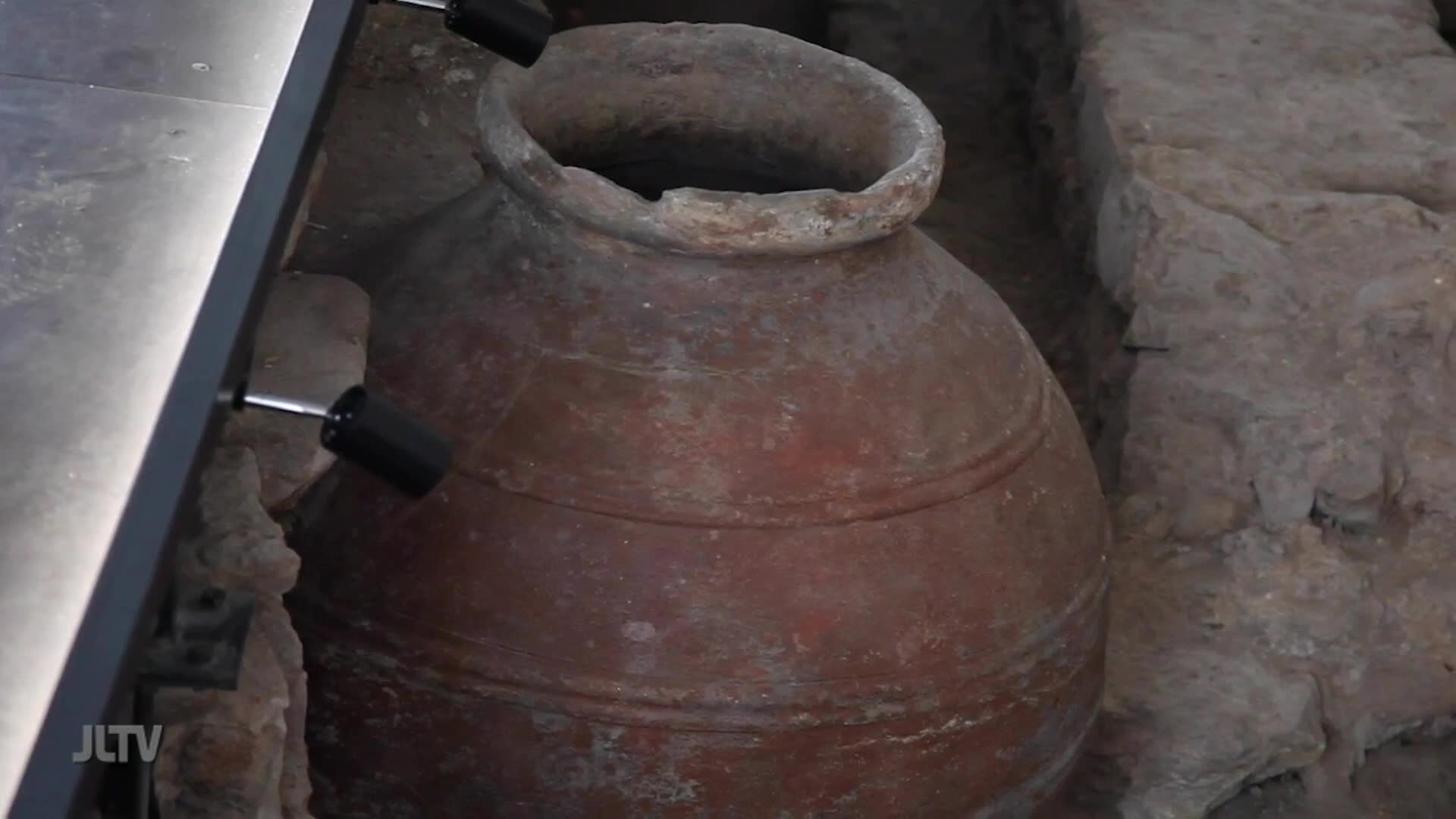 A large, ancient clay pot sits partially buried in the earth, its weathered surface showing rings of wear. Two small, black spotlights are positioned on a metal track, illuminating the artifact for display.