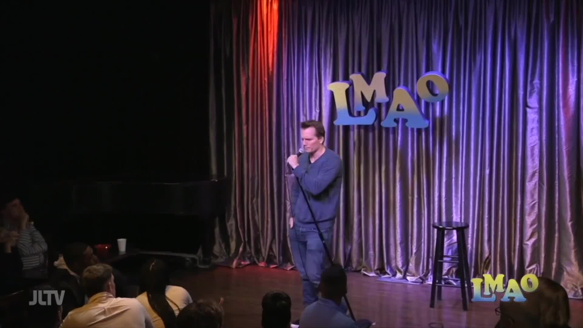 A comedian grips the microphone, bathed in stage lights against a shimmering curtain. The "LMAO" sign above him glows, a familiar sight for anyone watching Jewish Life Television in the United States.