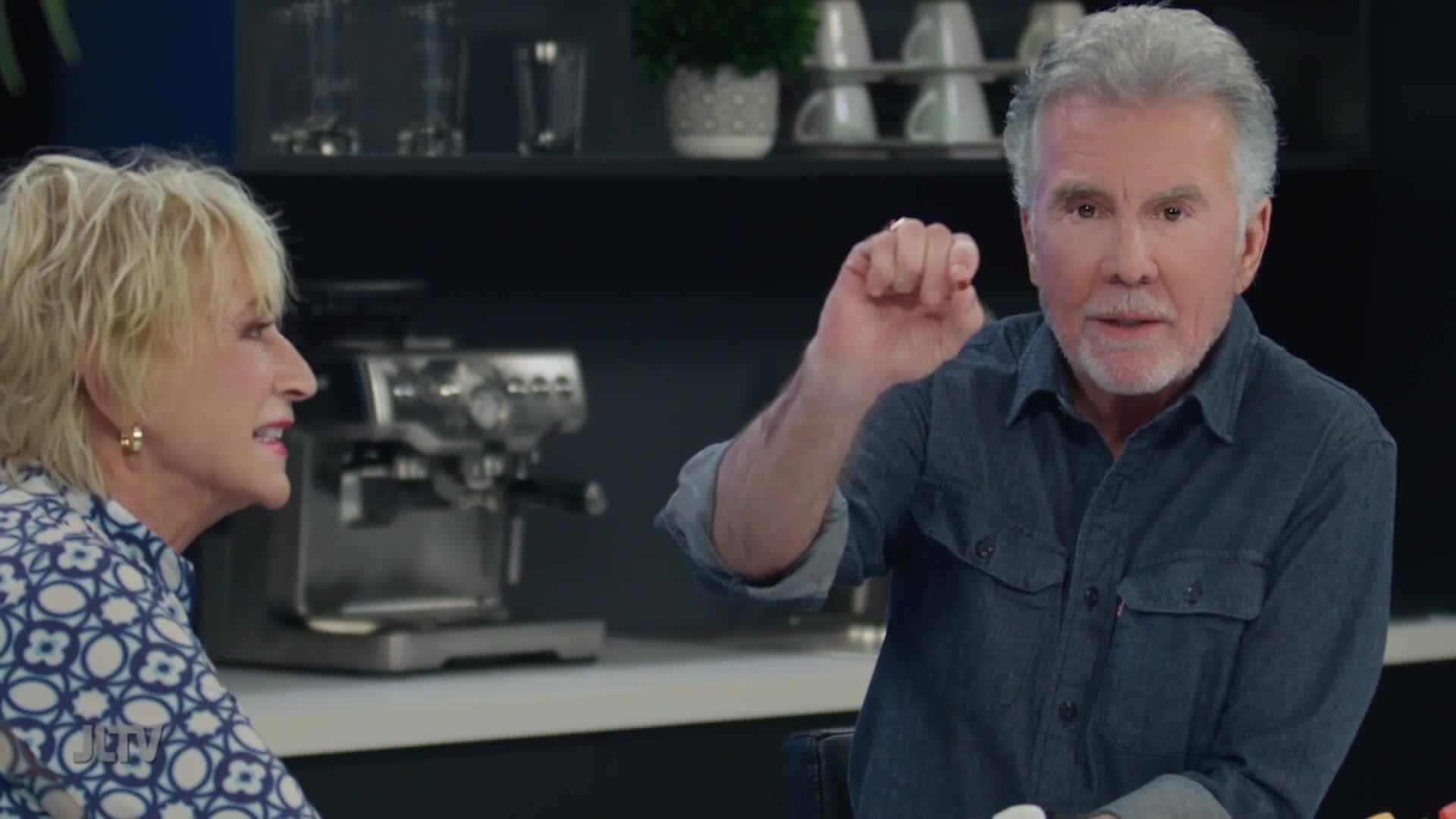 A man with grey hair gestures with his right hand, his index finger and thumb pinched together. Beside him, a woman with blonde hair turns her head to the side, her mouth slightly open.