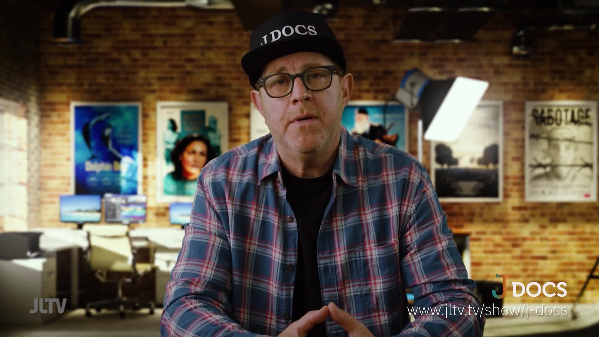 A man in a plaid shirt and a baseball cap sits in front of a brick wall. Posters hang behind him, and a bright light illuminates the scene.