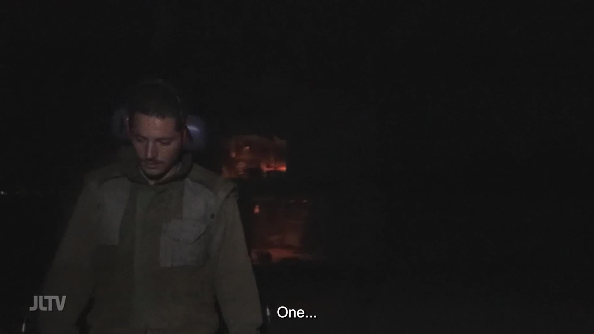 A young man in a military-style jacket walks through darkness, his face illuminated by a faint glow. Behind him, a building burns, its windows reflecting the flames.