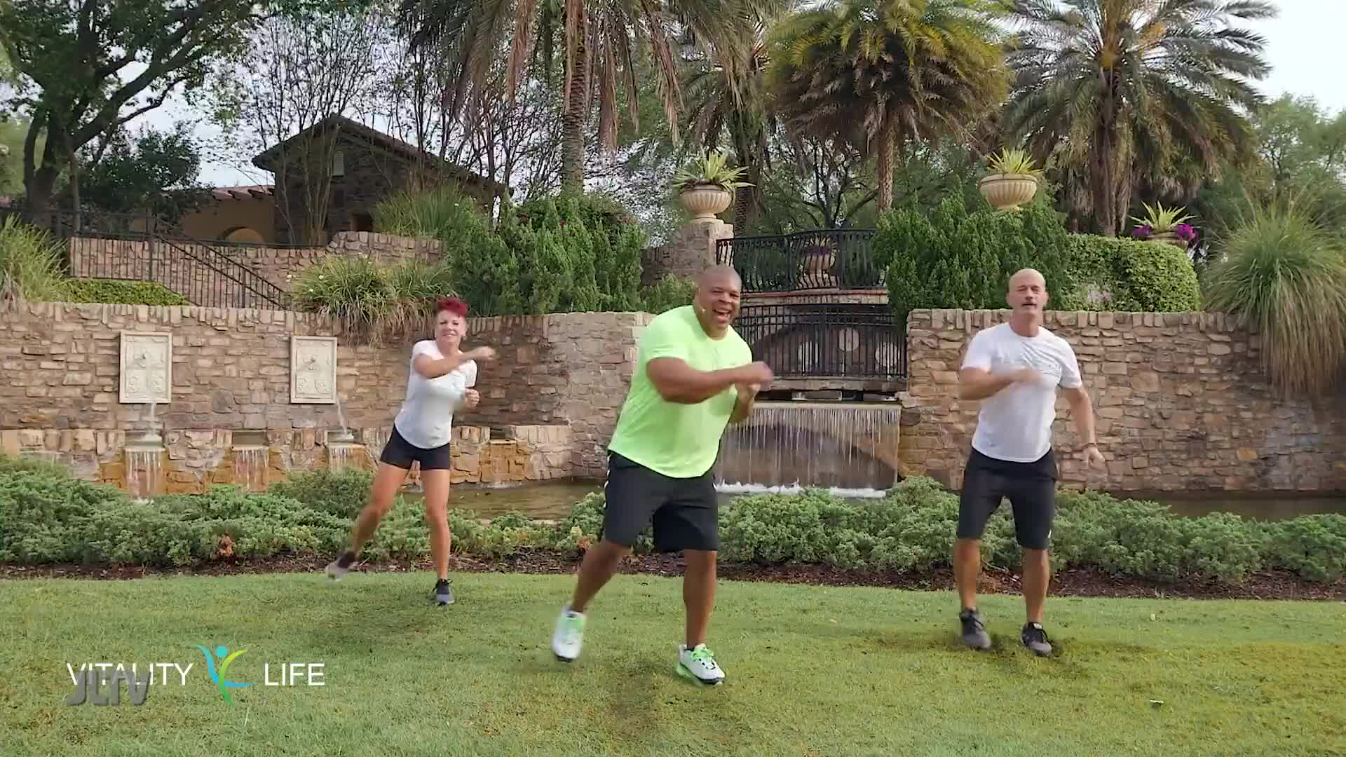 Three people are exercising outdoors on a grassy area with a stone wall and fountain behind them. The man in the bright green shirt is leading the movement, his arms bent and fists clenched.