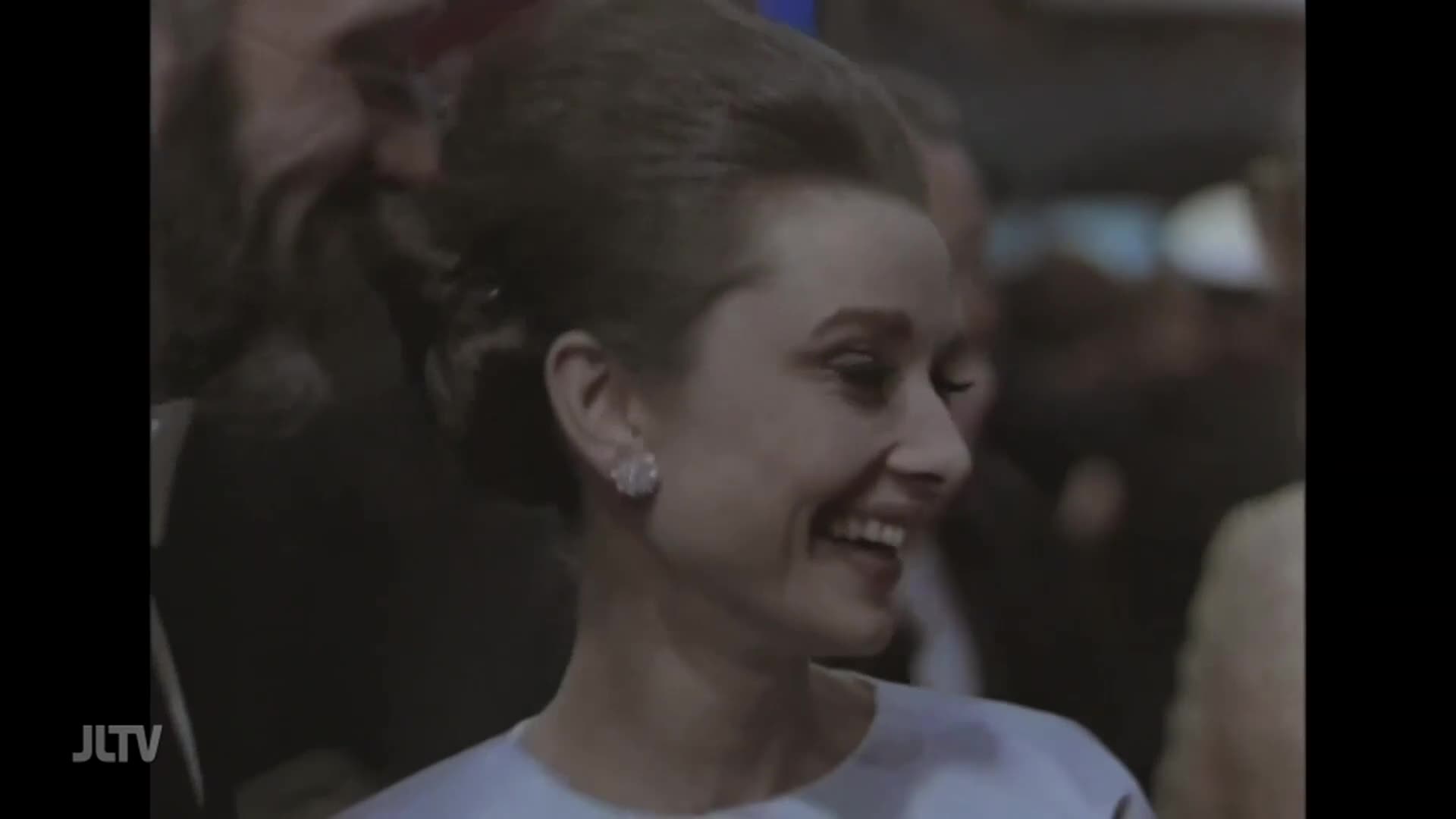 A woman with her hair swept up smiles broadly, her eyes crinkling. She's wearing a pale dress and sparkling earrings, surrounded by other people in the background.