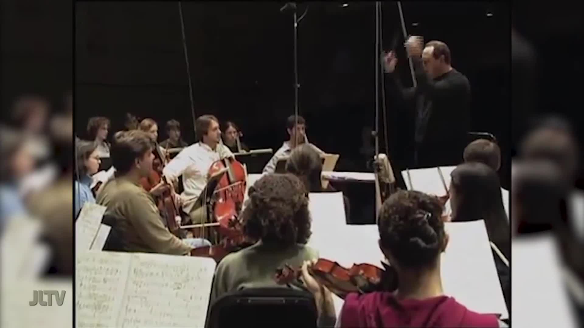 The conductor raises his hands, signaling the orchestra to begin. Cellos and violins are poised, ready to play.