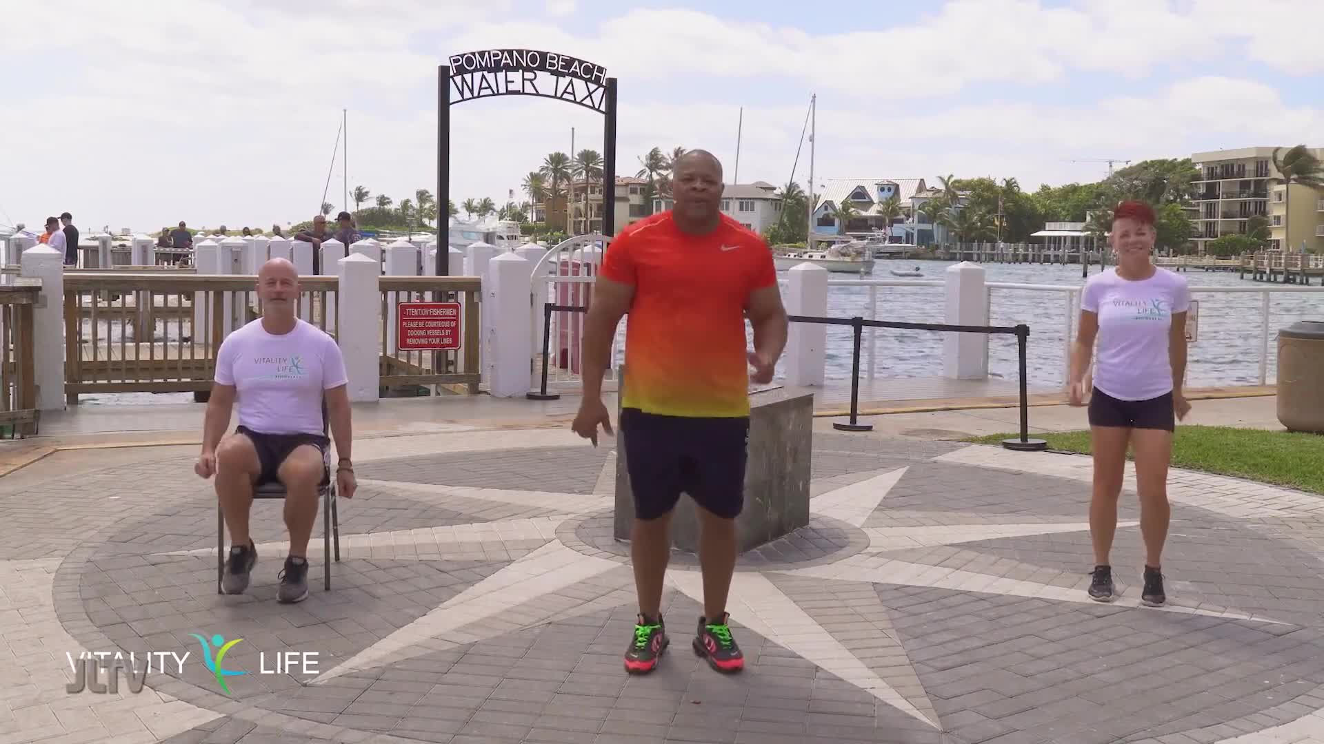 A man in an orange and red shirt stands center, arms bent at his sides, while a woman in a white t-shirt and shorts bounces lightly on her toes to his right. To the man's left, another man sits in a chair, his feet tapping the ground. This trio is exercising outdoors by a waterfront, with a sign for "Pompano Beach Water Taxi" visible behind them.