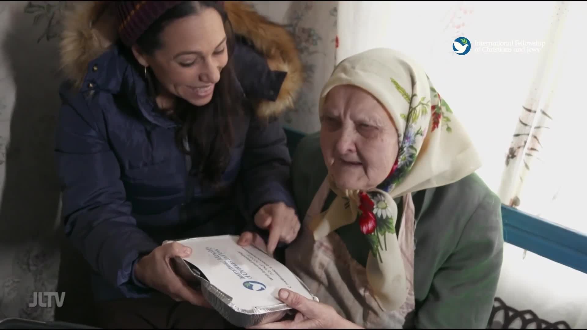 A woman in a blue puffer coat offers a foil-wrapped container to an elderly woman wearing a floral headscarf. The woman in the coat points to something on the container, her expression warm and engaged.