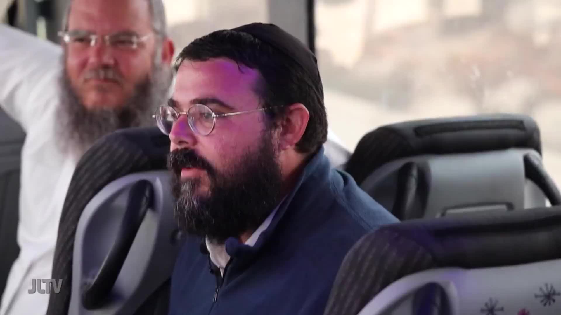 A man with a beard and glasses, wearing a kippah, sits in a bus seat. Another man, also bearded and wearing glasses, is visible behind him.
