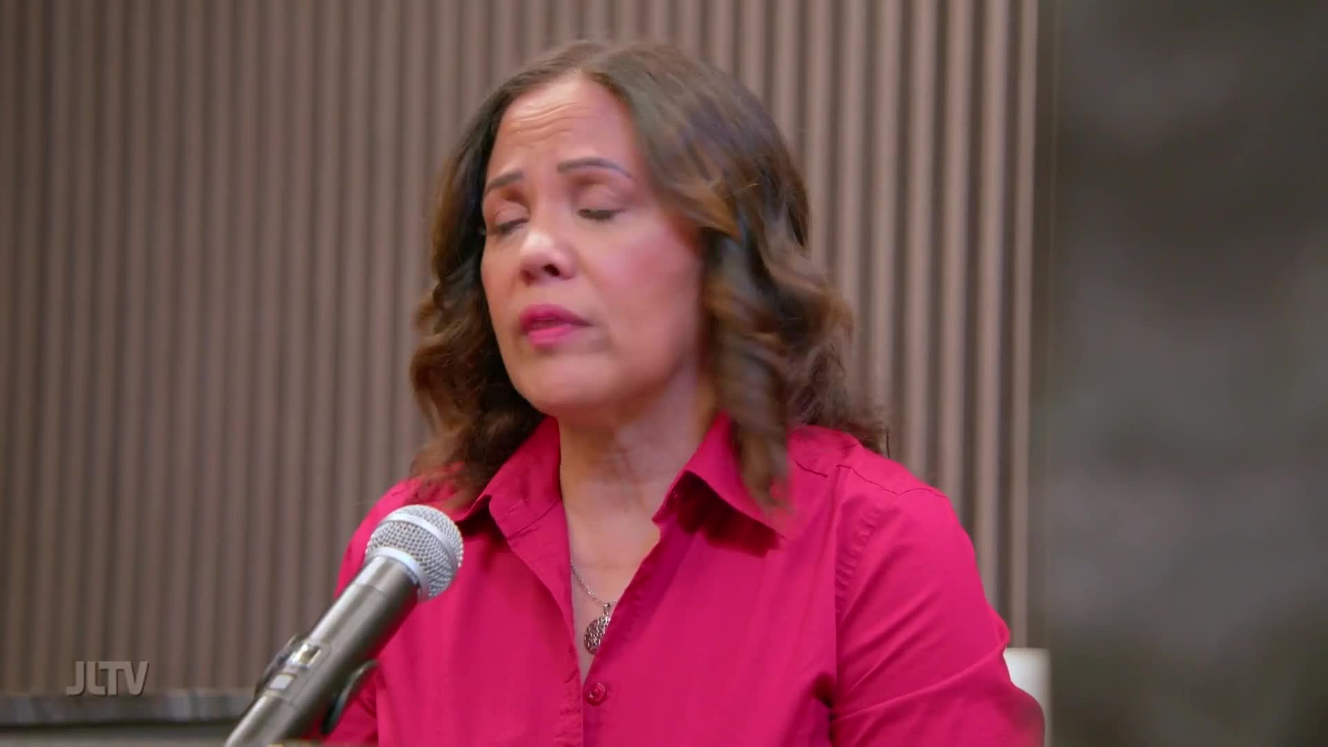 A woman in a bright pink shirt closes her eyes, her head tilted slightly as she speaks into a microphone. The JLTv logo appears in the corner of the screen.