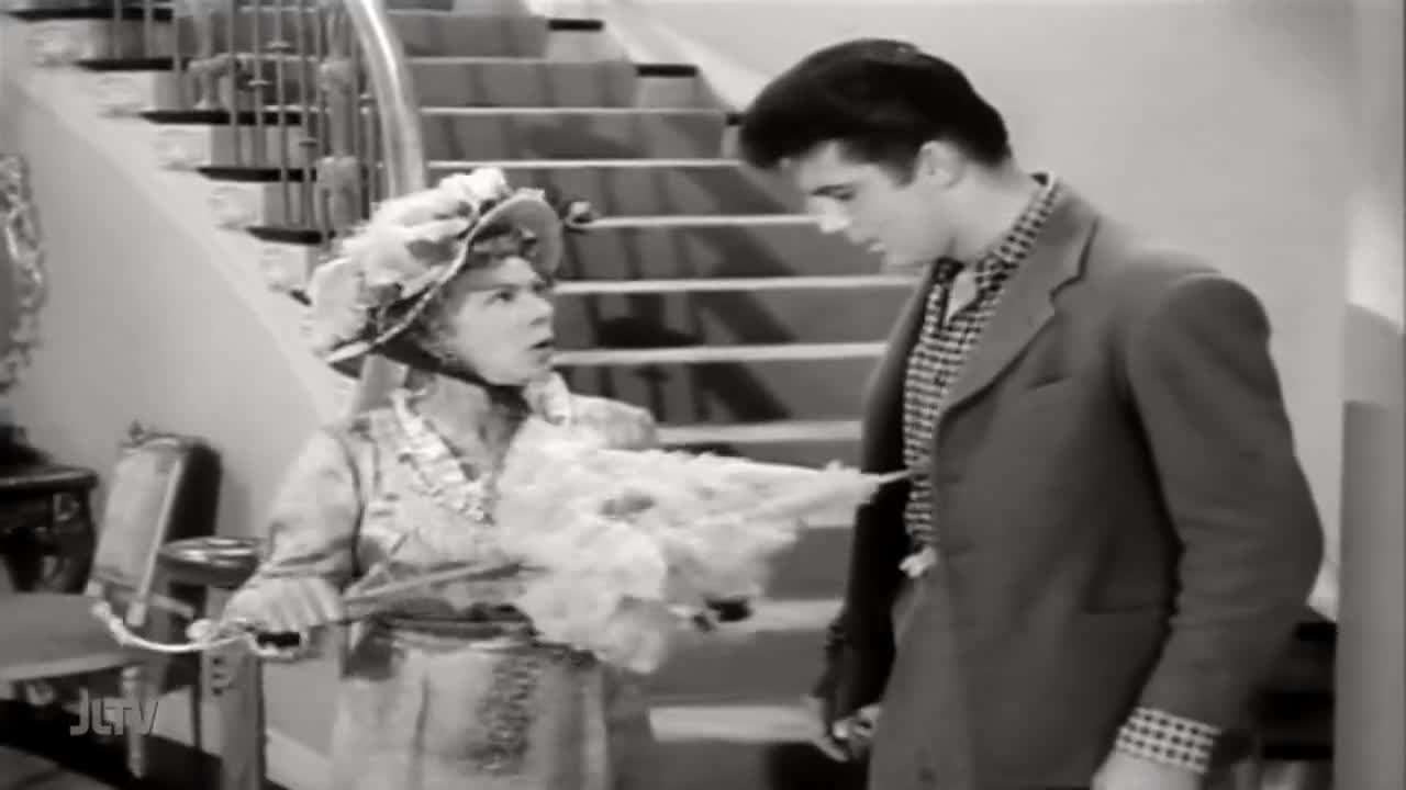 A woman in a wide-brimmed hat and a ruffled dress holds a feathered fan, her expression one of surprise as she speaks to a young man. He stands before her, dressed in a suit jacket over a patterned shirt, his gaze directed at her.