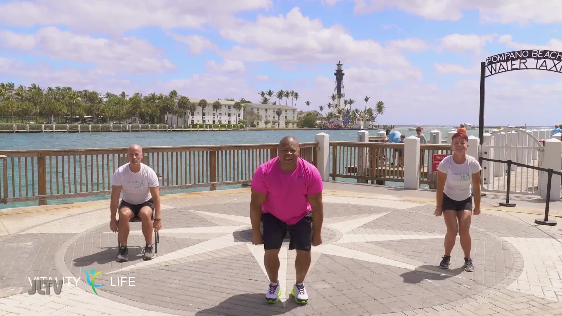 Three people are exercising outdoors on a sunny day by the water. A man in a bright pink shirt is in the center, with a man in white on his left and a woman in white on his right. The Pompano Beach Water Taxi sign is visible in the background.
