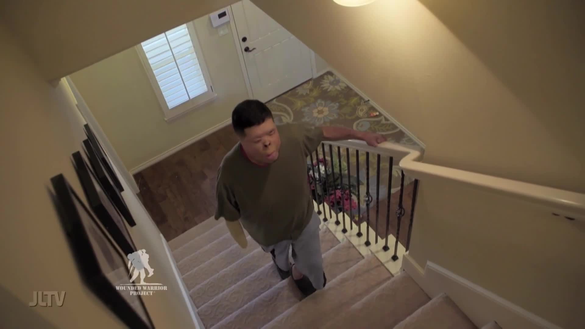 A man with a prosthetic arm ascends a carpeted staircase, his hand resting on the banister. The scene, captured for Jewish Life Television, highlights his determination.