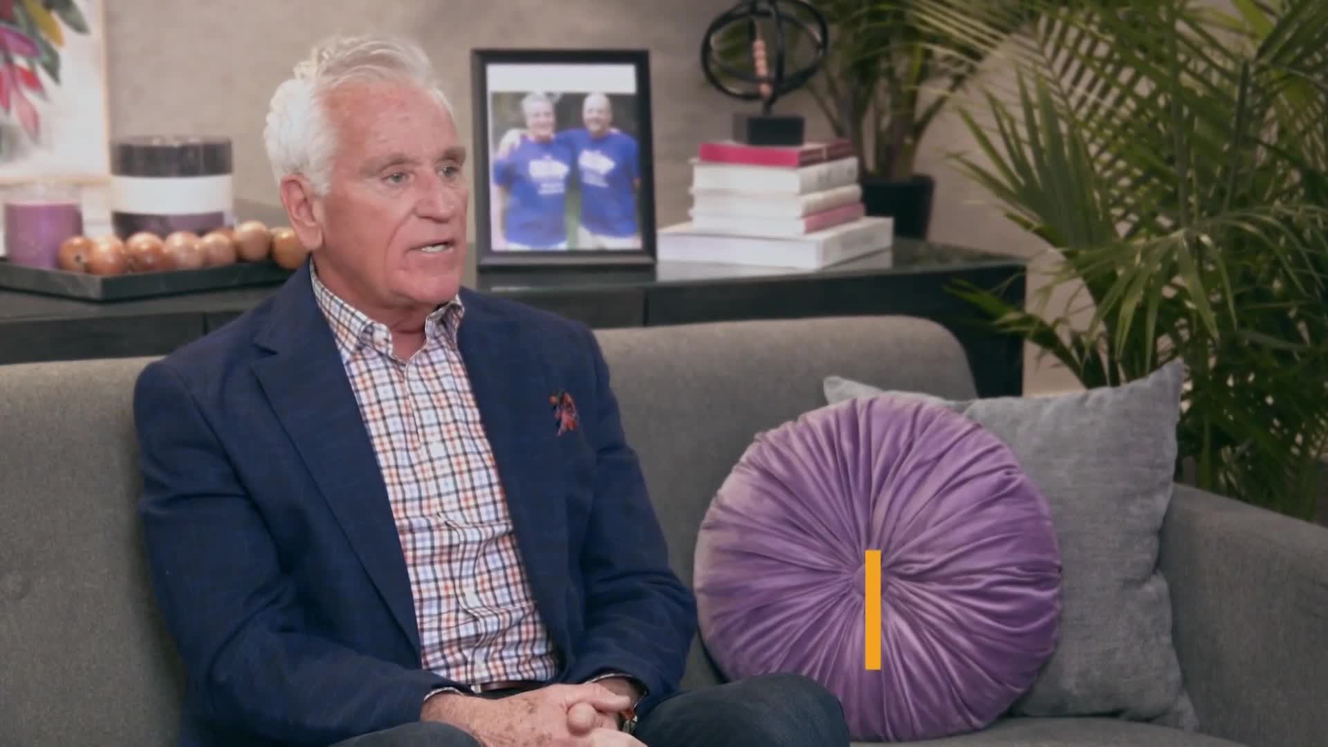 A man in a blue blazer and plaid shirt speaks from a couch, a framed photo of two people in matching shirts behind him. A plush purple cushion sits on the sofa next to him.