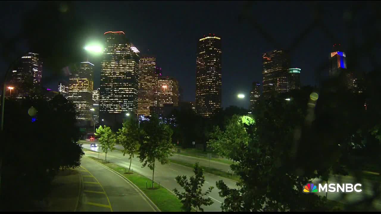 The Houston skyline glows with countless lit windows against the dark sky. Headlights of cars trace the curved road below, as seen on MSNBC.
