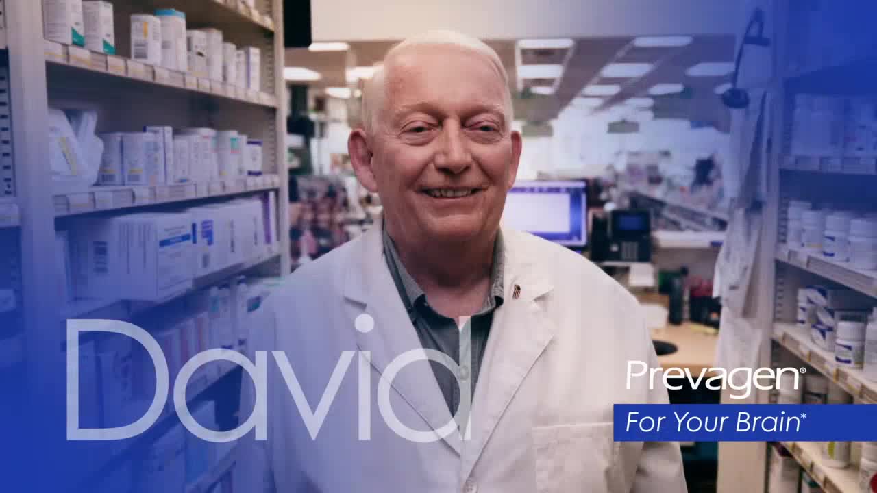 David, a pharmacist, smiles directly at the camera in a pharmacy setting. Shelves filled with medications line the background.
