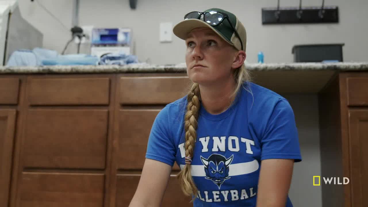 A woman in a baseball cap and a blue t-shirt looks up, her blonde braid hanging over her shoulder. The National Geographic Wild logo is visible in the corner.
