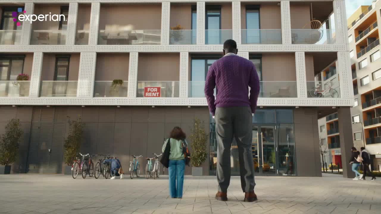 A man in a purple sweater stands with his back to me, looking up at a modern apartment building. A young girl walks past him, heading towards the entrance.
