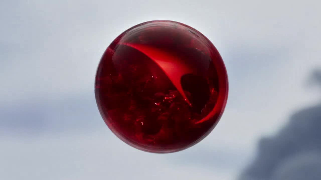 A perfectly round, translucent red sphere floats against a pale sky. Within the orb, a bright, curved streak of light dances across its surface.
