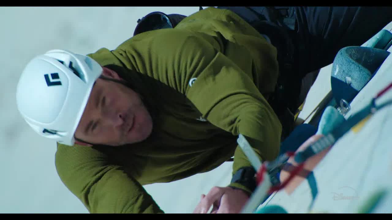 He's gripping the rock face with one hand, his other hand reaching for a hold. His white helmet and green shirt stand out against the pale rock.
