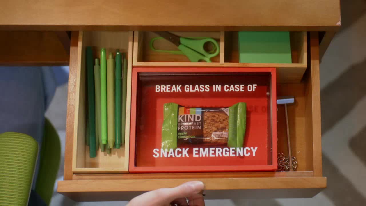 A wooden drawer slides open, revealing a "Snack Emergency" kit. Inside, a KIND bar sits behind a red frame with the words "Break Glass In Case Of" printed above it.
