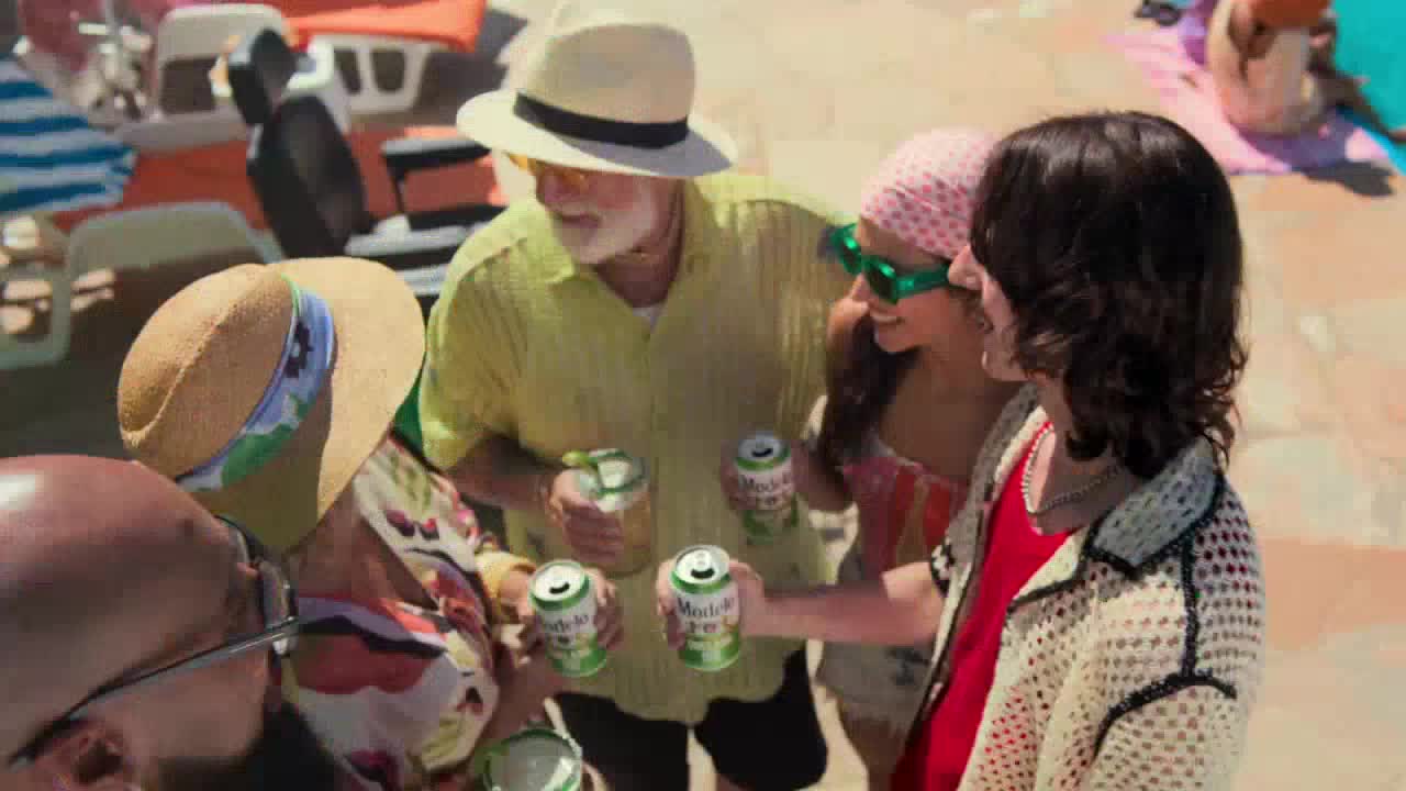 A group of people are gathered, holding green cans, likely beer, near a pool. The man in the center, wearing a straw hat, is smiling as he looks at the others.

