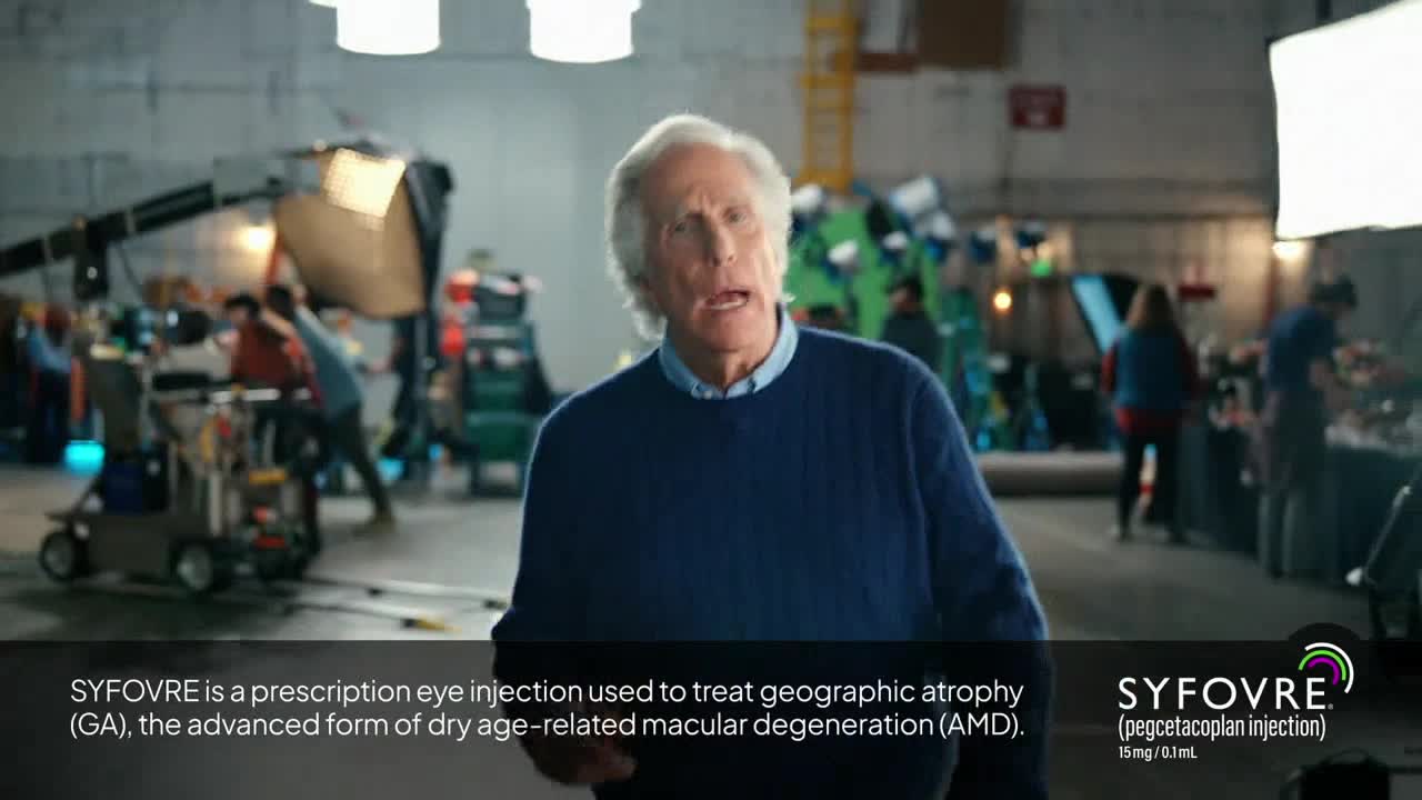 A man in a blue sweater speaks directly to the camera, while crew members mill about in the background. The set is brightly lit, and a logo for a prescription eye injection appears below.

