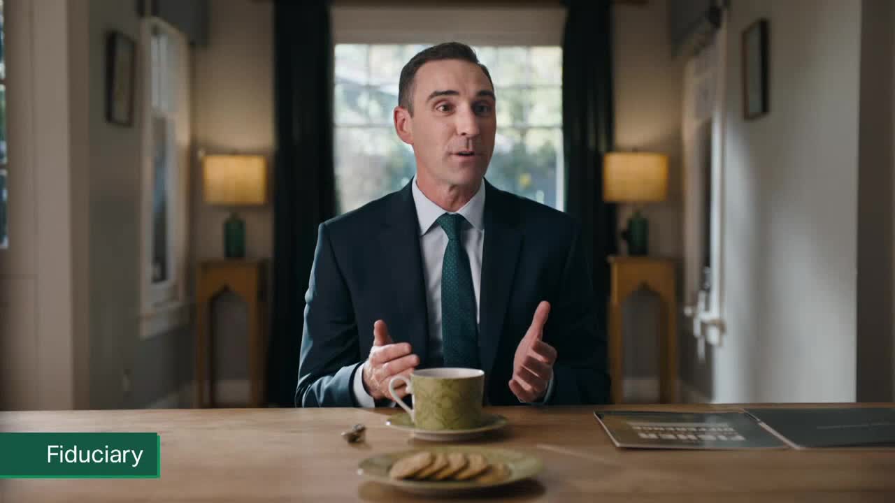 A man in a dark suit gestures with his hands, speaking to someone off-camera. On the table in front of him are a teacup, a saucer with cookies, and a few documents.
