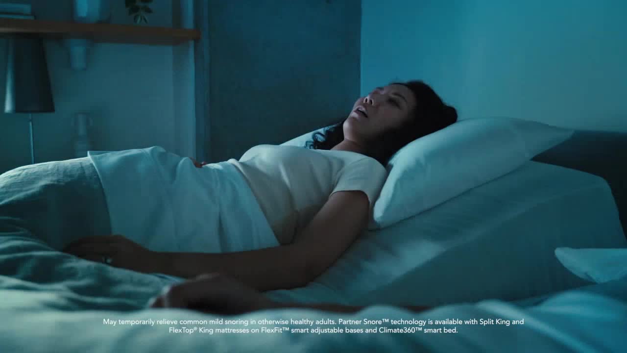 A woman lies in bed, her mouth slightly open as if she's breathing heavily. The scene is dimly lit, suggesting it is nighttime.
