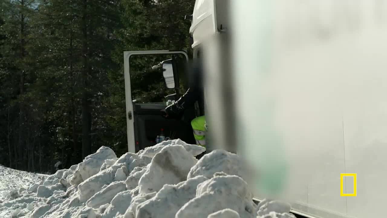 A person in dark clothing is getting out of a white truck parked beside a snowbank. The open door reveals a glimpse of a bright green vest.
