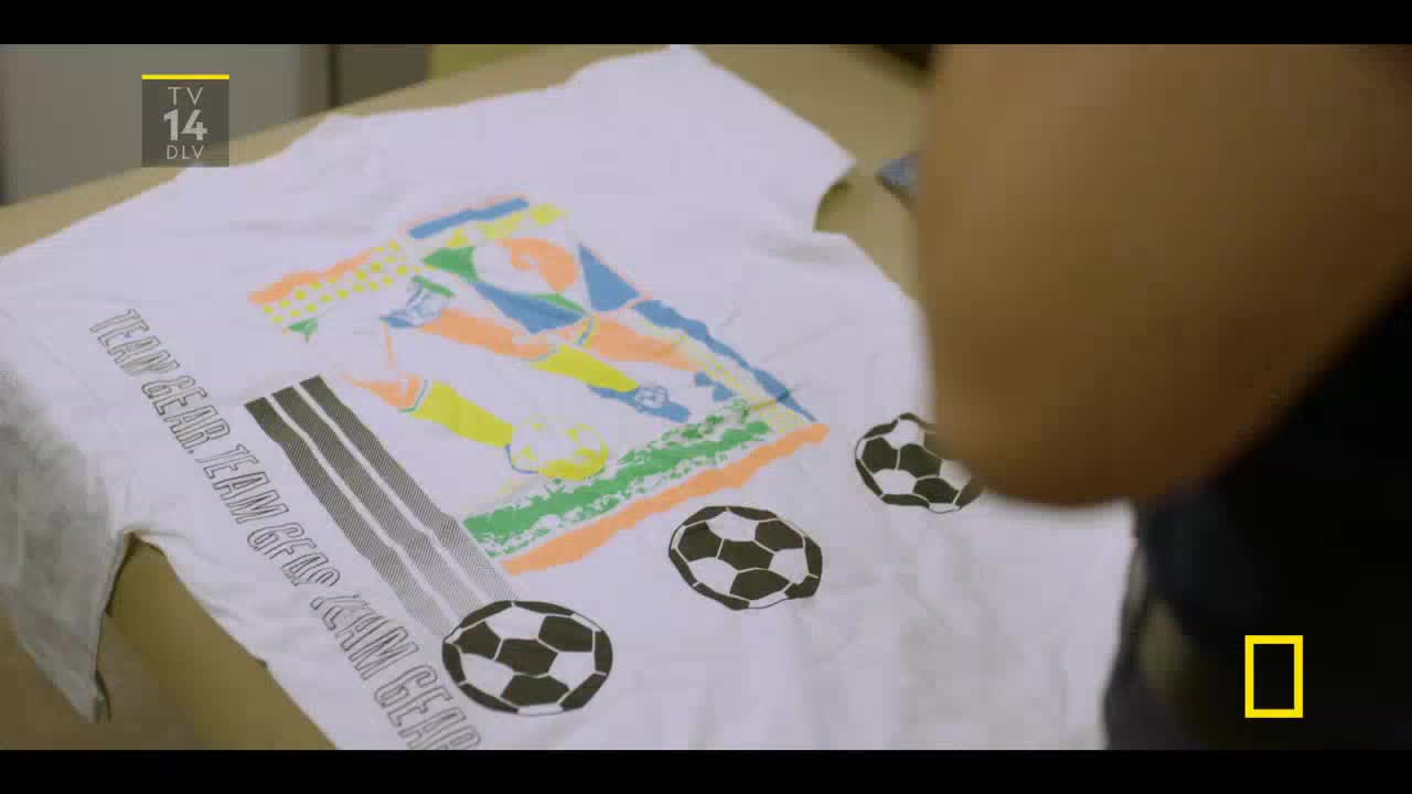 A white t-shirt, emblazoned with soccer balls and a player graphic, lies on a table. Someone's arm is in the frame, likely reaching for the shirt, as a National Geographic logo appears in the corner.
