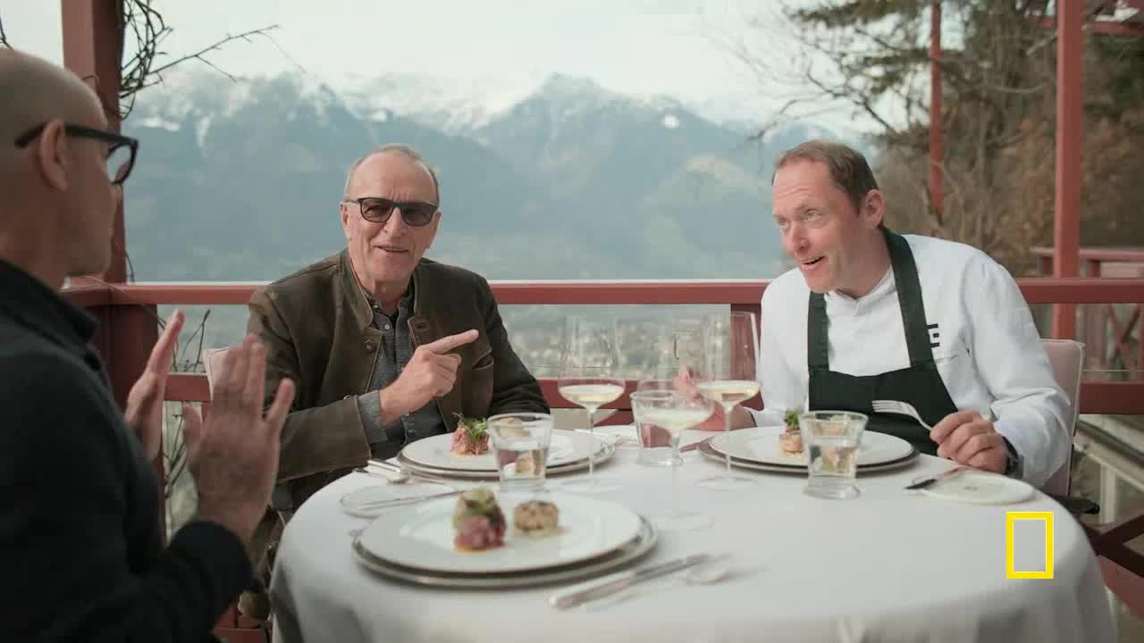 Three men are seated around a white tablecloth, gesturing animatedly at each other. One man in a chef's coat leans forward, while another points to the food on the table.
