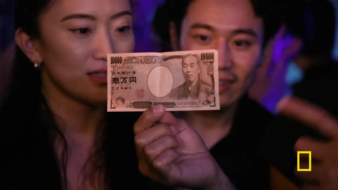 A man holds up a Japanese 10,000 yen note, showing it to a woman beside him. The bill features a portrait and intricate script, illuminated by the ambient light of a dimly lit space.
