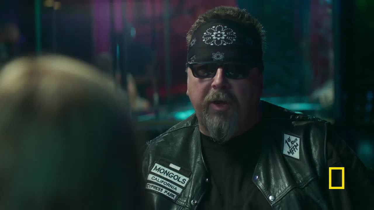 A man in a leather vest, bandana, and sunglasses is speaking, his mouth open. The National Geographic logo is visible in the corner, and the "Mongols" patch is clear on his vest.
