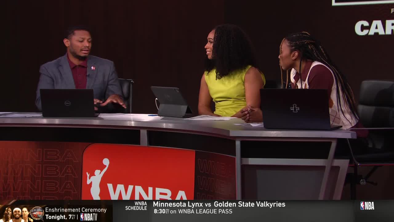 Three people are seated at a desk, each with a laptop in front of them. The man in the center gestures with his hands while the women on either side listen intently.
