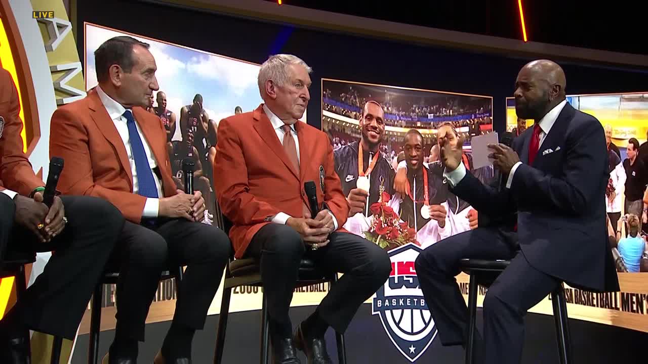 Three men in orange blazers sit on stools, listening to a man in a suit who holds a small card. Behind them, a backdrop displays a photo of basketball players with medals.

