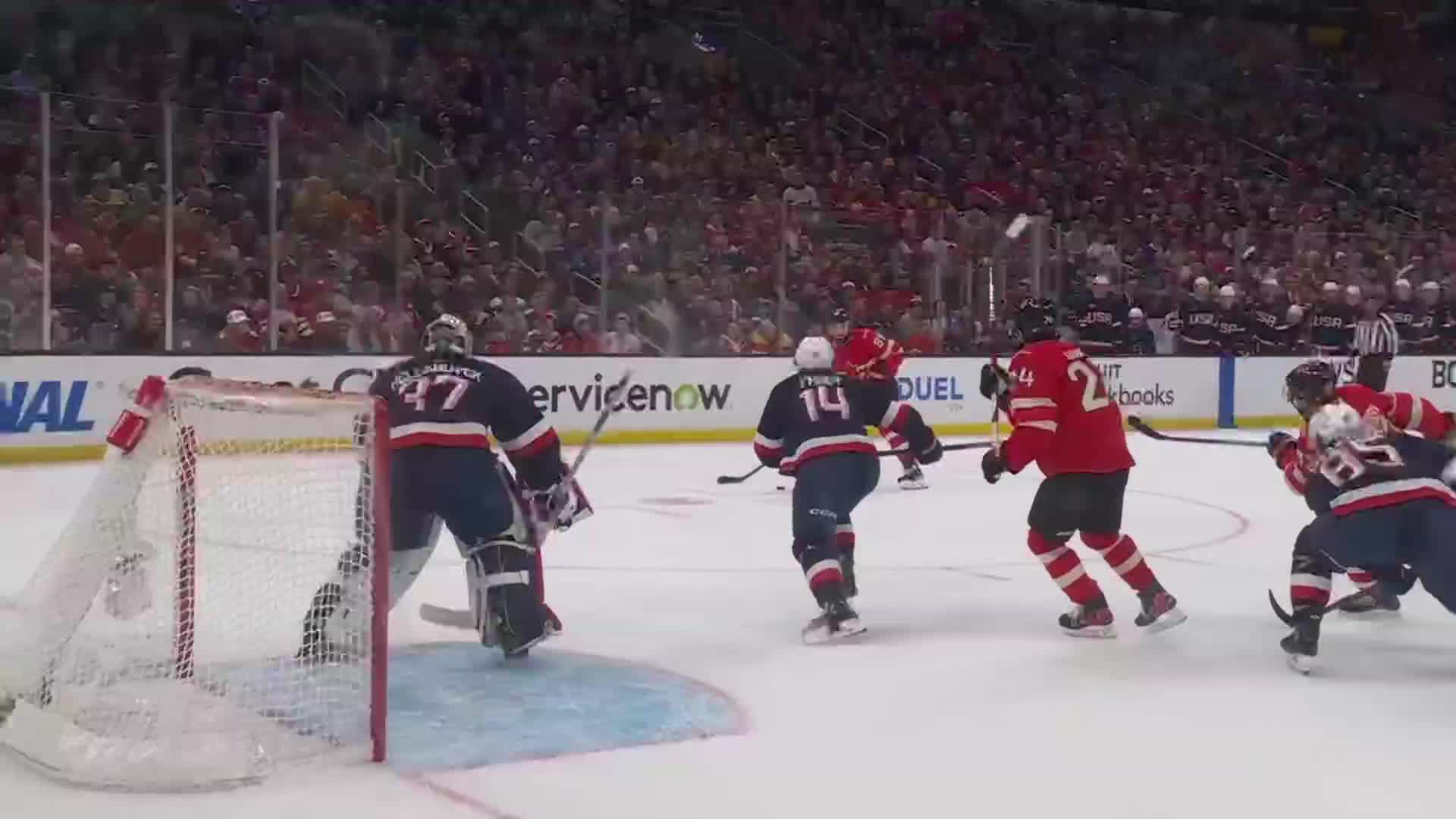 A player in a red jersey with the number 24 skates towards the net, while a goalie in navy and red gear prepares to make a save. The puck is in the air near the net.
